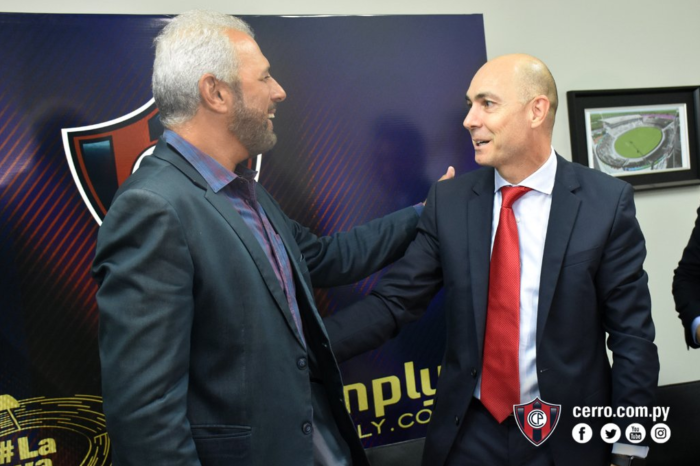 Dois homens sorrindo e interagindo em frente a um painel com os logos do Cerro Porteño (CP), "imply", a hashtag "#LaNuevaOlla" e, à direita, o site "cerro.com.py" com ícones de redes sociais. Um quadro com a foto de um estádio está ao fundo.