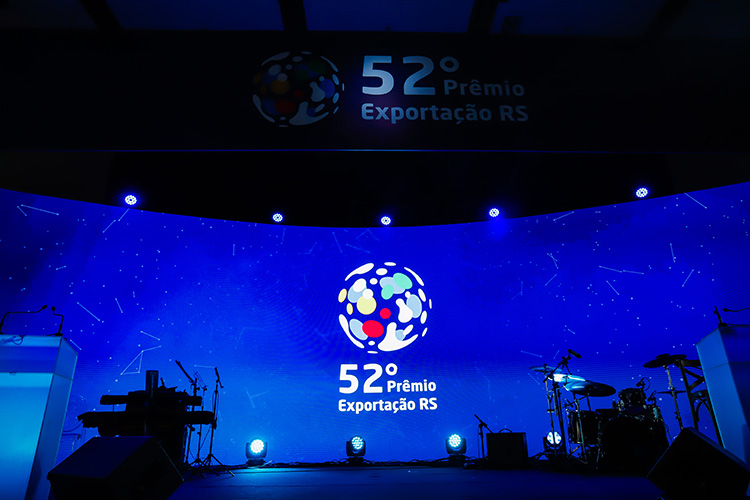 Um palco iluminado em azul com um grande telão exibindo o logotipo do "52º Prêmio Exportação RS". Há instrumentos musicais e equipamentos de som no palco.