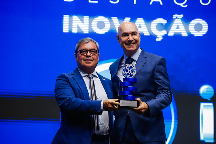 Dois homens em ternos azuis recebem um prêmio de inovação em um palco com um fundo azul. O troféu é composto por anéis azuis empilhados sobre uma base metálica.
