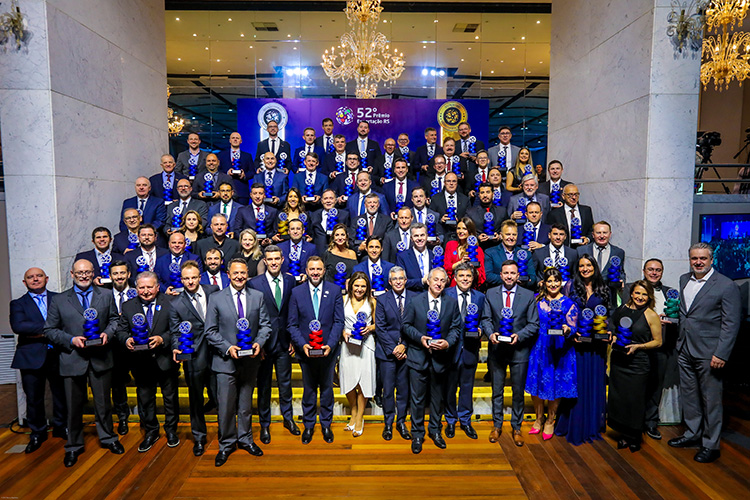 Grande grupo de pessoas em trajes formais, segurando troféus azuis e dourados, posam para uma foto em um evento de premiação.