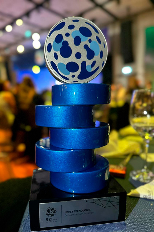 Prêmio azul em camadas com topo em forma de globo prateado e azul-escuro. Base preta com placa identificando o prêmio como "Imply Tecnologia".