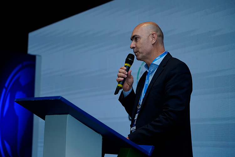 Um homem careca de terno escuro fala em um púlpito, segurando um microfone sem fio. Um painel azul está visível ao fundo.