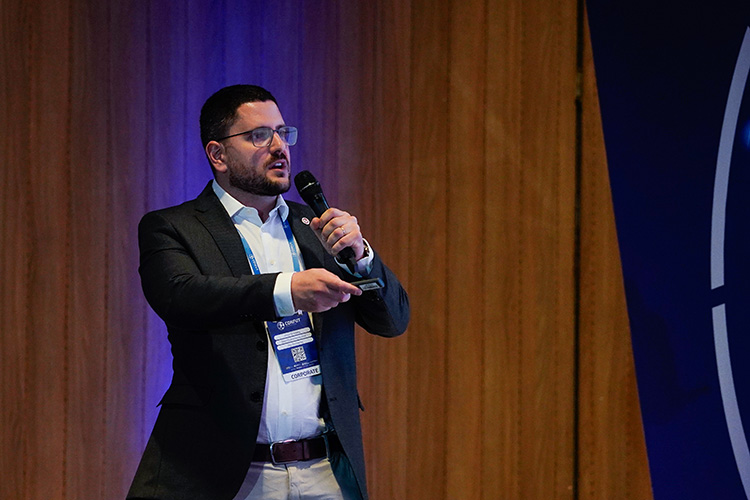 Homem de terno escuro falando em um microfone em um evento corporativo. Ele usa óculos e está gesticulando.