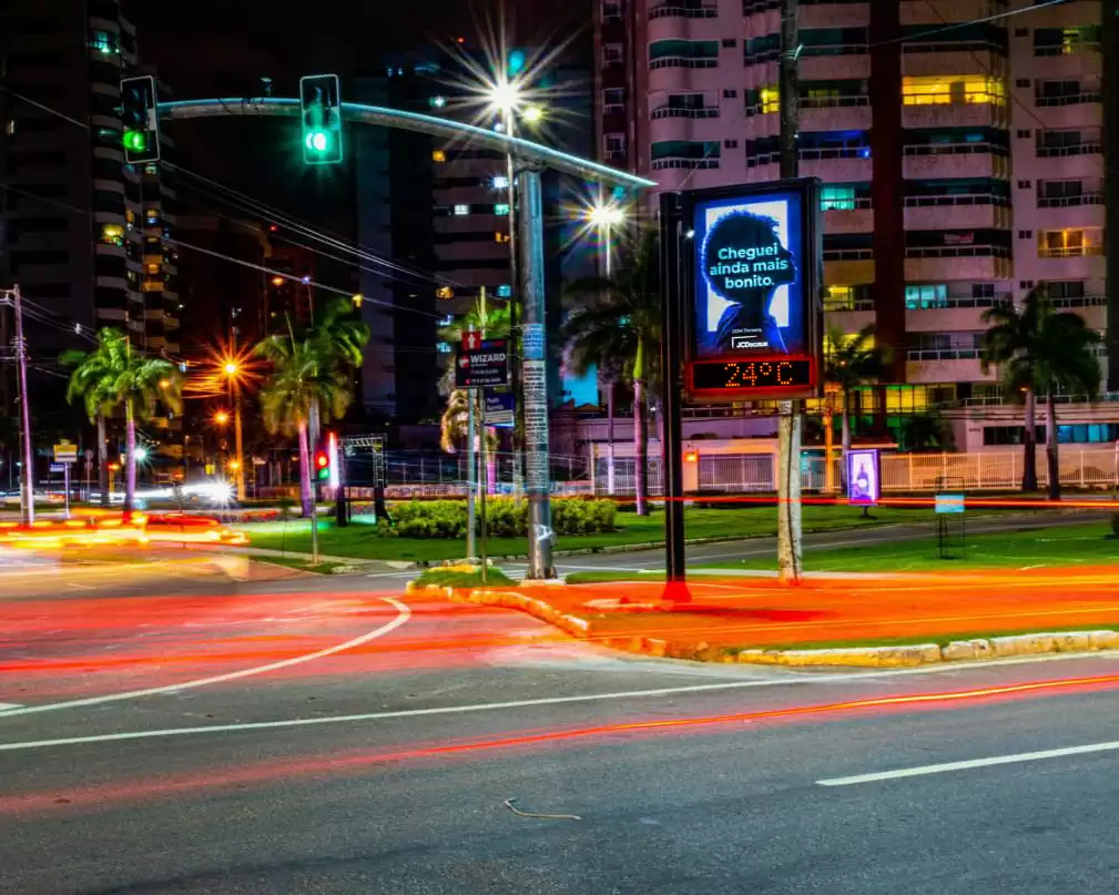 Foto de uma rua com um semáforo e ao lado um Relógio Digital e ao fundo alguns predios.