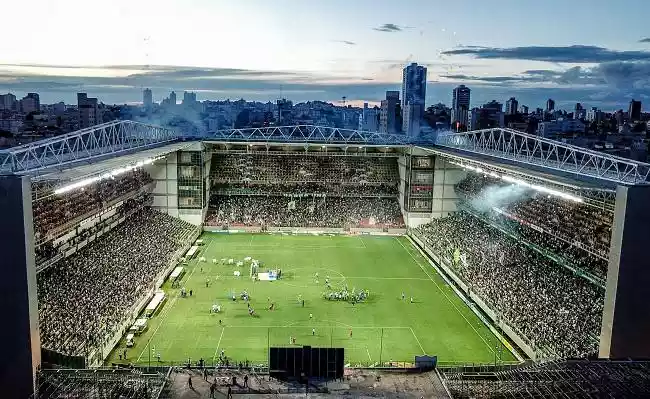 Estádio lotado à noite, com campo de futebol iluminado e cidade ao fundo. Muitas pessoas na arquibancada.