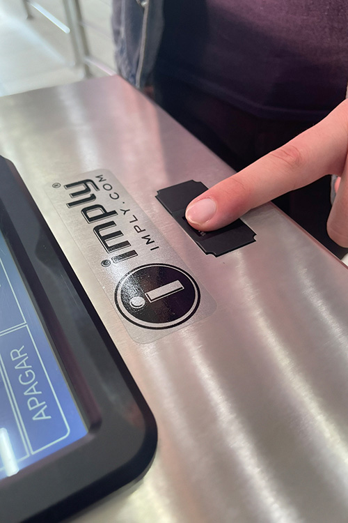 Um dedo pressionando um leitor biométrico Imply em uma superfície de aço inoxidável. O logotipo da Imply está visível na superfície.