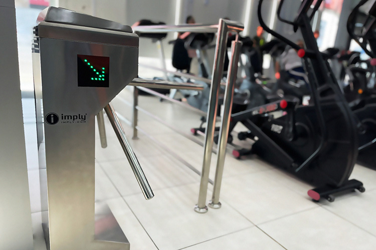 Torniquete de acesso eletrônico de metal prateado com uma seta verde indicando a direção, em uma academia com equipamentos de exercícios ao fundo.