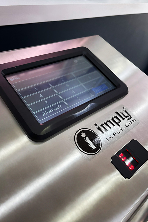 Close-up de um painel de controle de aço inoxidável com teclado numérico, leitor de impressão digital e logo da Imply. A tela mostra um teclado numérico com a palavra "Apagar".