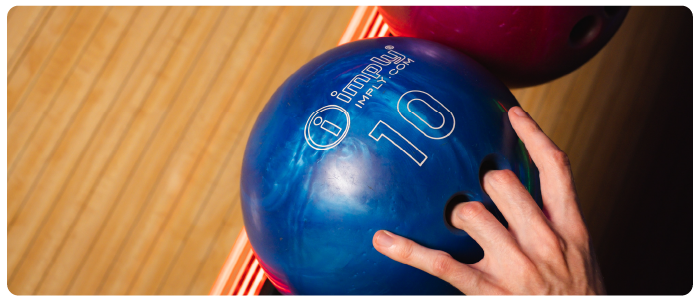 Uma pessoa com os dedos médio e anelar nos buracos de uma bola de boliche azul escrito "imply - imply.com -10".