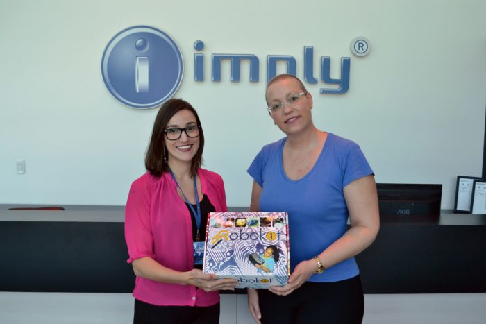 Duas mulheres sorrindo enquanto seguram uma caixa do jogo BOKA na recepção da empresa Imply, com o logotipo da empresa ao fundo.