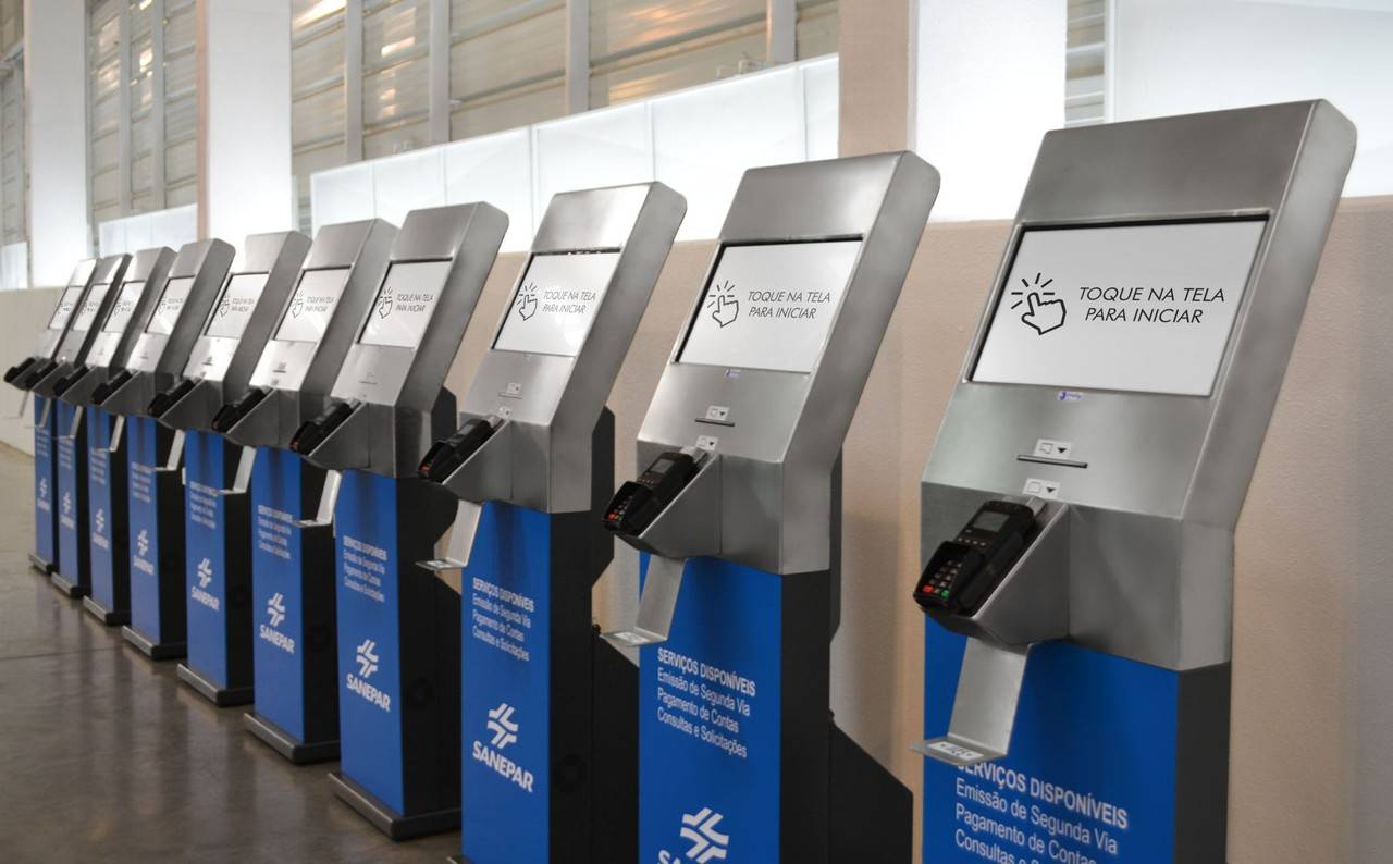 Fila de terminais de autoatendimento da Sanepar com telas exibindo a mensagem 'Toque na tela para iniciar' e maquininhas de pagamento acopladas.