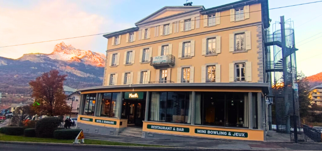 Um prédio de três andares de cor creme com um restaurante e bar no térreo, com uma escada em espiral externa e montanhas ao fundo.