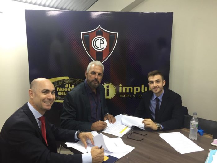 Três homens de terno sentados à mesa assinando documentos, diante de um painel com o escudo do Cerro Porteño (CP), o logo da "imply" e a hashtag "#LaNuevaOlla".