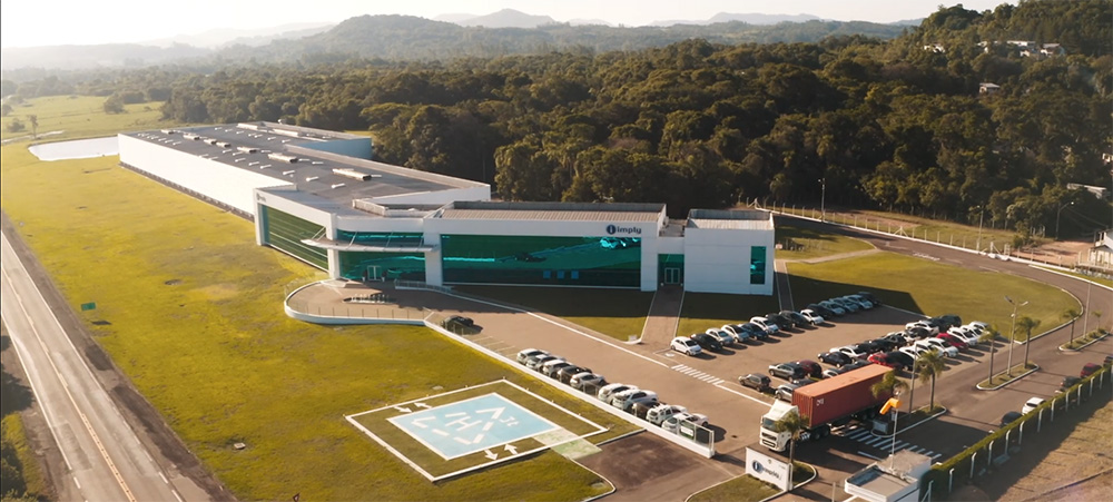 Visão externa da sede da Imply em Santa Cruz do Sul. Mostra a fachada e toda sua ampla estrutura, incluindo o lado ao fundo e o bosque ao redor que faz parte da empresa.