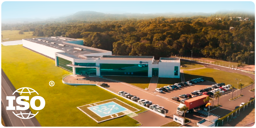 Vista aérea de uma fábrica moderna e extensa, cercada por vegetação, com estacionamento e um heliponto. O logotipo da ISO está visível no canto inferior esquerdo.