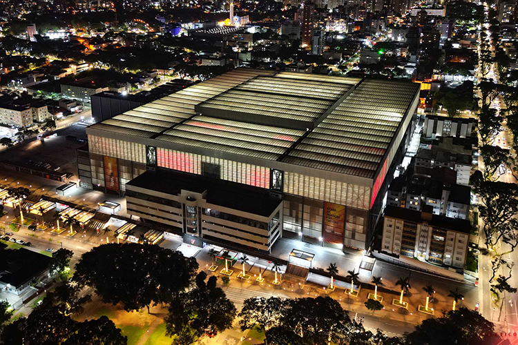 Vista aérea noturna de um grande complexo de edifícios iluminado, com telhado de estrutura de vidro e rodeado por árvores e ruas da cidade.