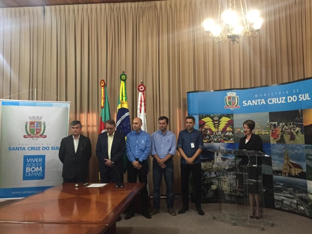 Fotografia de um evento oficial no município de Santa Cruz do Sul, com seis homens e uma mulher em pé ao lado de uma mesa. No fundo, há bandeiras do Brasil, do estado do Rio Grande do Sul e do município, além de banners institucionais com o brasão e imagens da cidade. A mulher está em um púlpito de acrílico, aparentemente discursando, enquanto os demais estão perfilados ao lado. A sala tem cortinas bege e um lustre iluminado no teto.