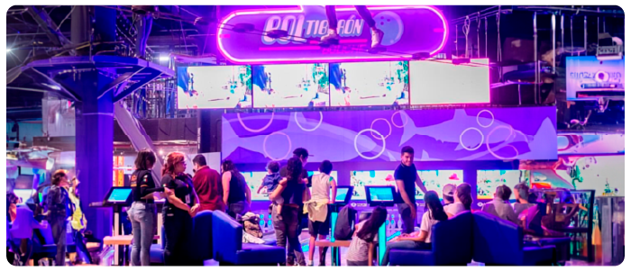 Imagem de um centro de entretenimento com iluminação neon em tons de roxo e azul. No topo, há uma placa iluminada com o nome "BOL TIB-FUN" e um ícone de bola de boliche. Abaixo da placa, várias telas exibem imagens de jogos ou vídeos. O ambiente está cheio de pessoas interagindo com telas sensíveis ao toque, sentadas em sofás ou caminhando pelo local. O fundo apresenta um design com círculos e montanhas estilizadas, reforçando o clima futurista e dinâmico do espaço.