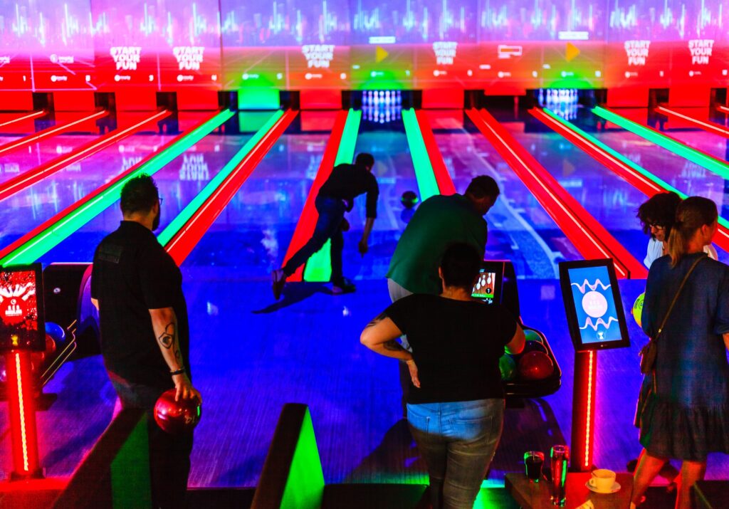 Pessoas jogando boliche em uma pista iluminada com luzes neon vermelhas e verdes. As telas de fundo mostram a frase "Comece sua diversão".