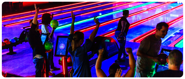 Várias pessoas em uma pista de boliche com as mãos levantadas ao lado dos totens de autoatendimento e dos repositores de bola.