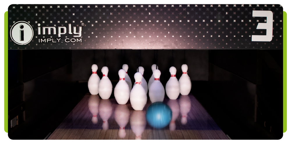 O final de uma pista de boliche com os dez pinos levantados prestes a serem derrubados por uma bola de boliche, na parte de cima temos o nome e logo da empresa "imply - imply.com" e o número "três". 