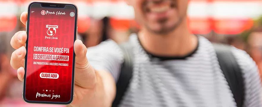 Uma pessoa segura um smartphone mostrando uma tela vermelha com a mensagem: "Confira se você foi o sorteado a ganhar um produto das coleções passadas".  Há um botão "Clique aqui" e a menção de "Próximos jogos".