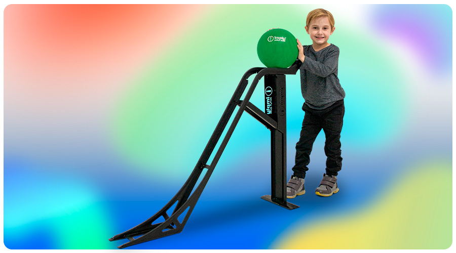 Criança em pé com as mãos em uma bola de boliche, que está em um suporte para escorregar a bola, ao fundo há uma mistura de cores em efeito embaçado.