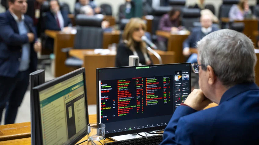 Homem sentado visualizando dois monitores, ao fundo há várias pessoas sentadas.
