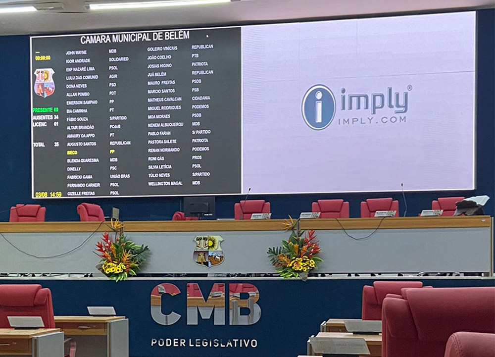 Fotografia do plenário da Câmara Municipal de Belém, com um painel eletrônico exibindo a lista de parlamentares presentes, ausentes e licenciados. O ambiente possui cadeiras vermelhas e uma mesa diretora decorada com arranjos florais. No painel, além da lista de nomes e partidos, há a logomarca da empresa 'Imply'. A sigla 'CMB' e a inscrição 'Poder Legislativo' estão visíveis na parte frontal do plenário.