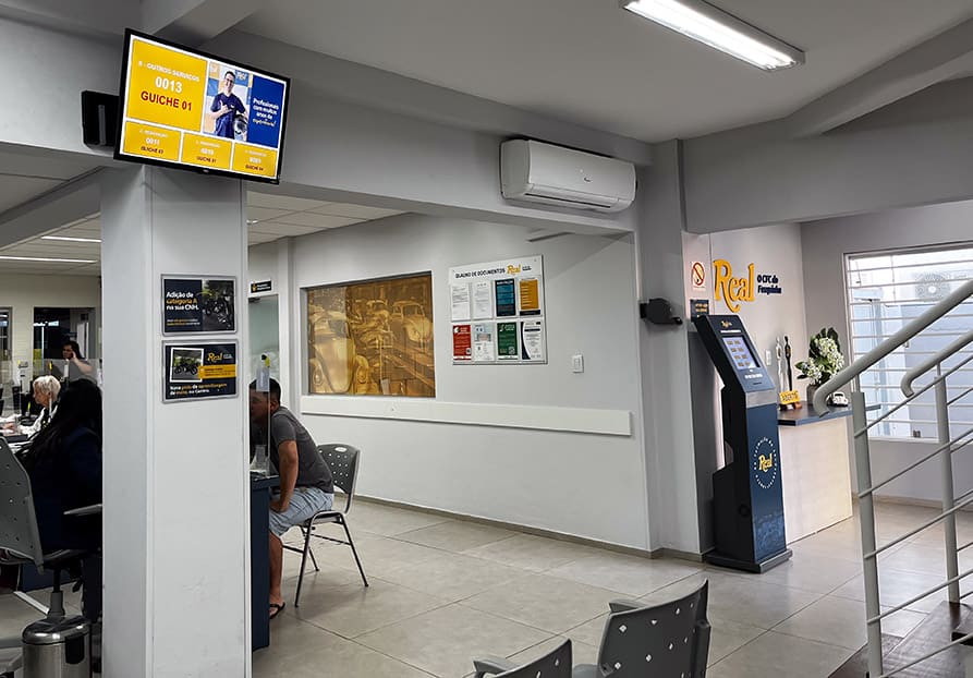 Sala de espera com painel de senhas de atendimento em cima de uma coluna
