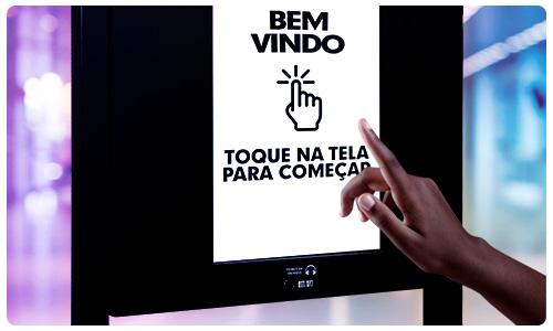 Um totem de autoatendimento com a frase escrita na tela: "BEM VINDO" e "TOQUE NA TELA PARA COMEÇAR"